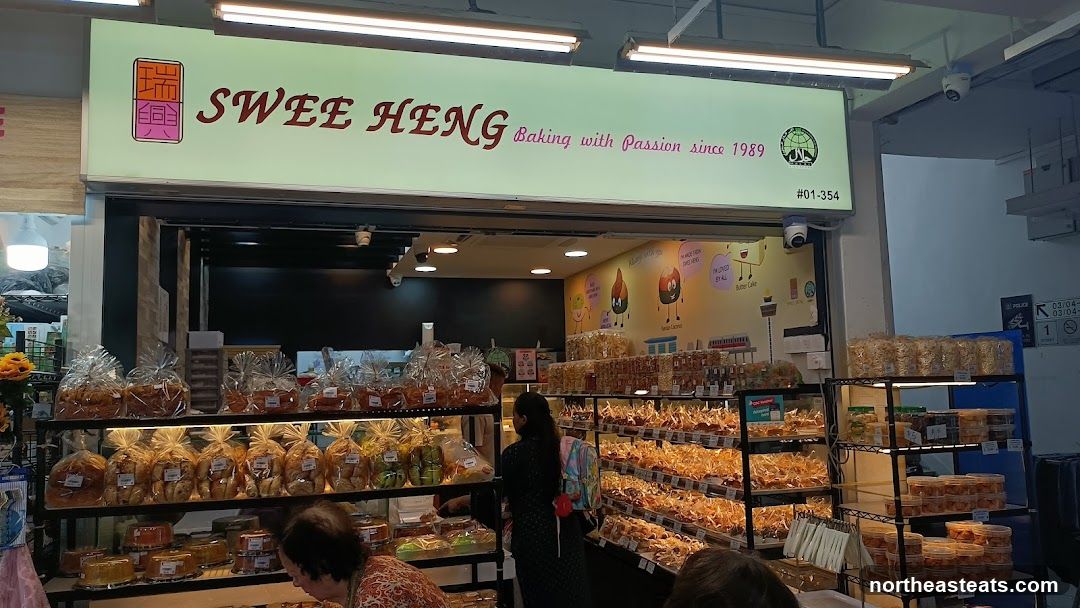 A bakery storefront with a large sign that reads "SWEE HENG Baking with Passion since 1989". The interior is visible, displaying shelves filled with various baked goods packaged in plastic bags and containers. Several customers are browsing the products.