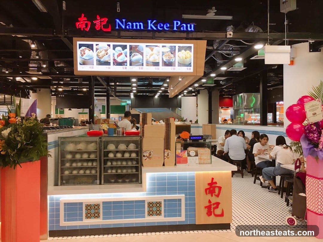 A food stall with a prominent sign that reads "Nam Kee Pau" in English and Chinese characters. Above the counter, a digital menu displays images of food items with prices. The counter itself has glass display cases filled with steamed buns. Customers are seated at tables in the background.