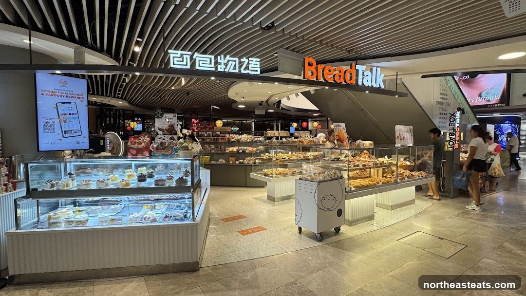 A BreadTalk bakery storefront with display cases filled with cakes and pastries. The store features prominent signage with the BreadTalk logo and Chinese characters. There are customers browsing the selection and staff attending to them. The interior is well-lit with modern shelving and display units.