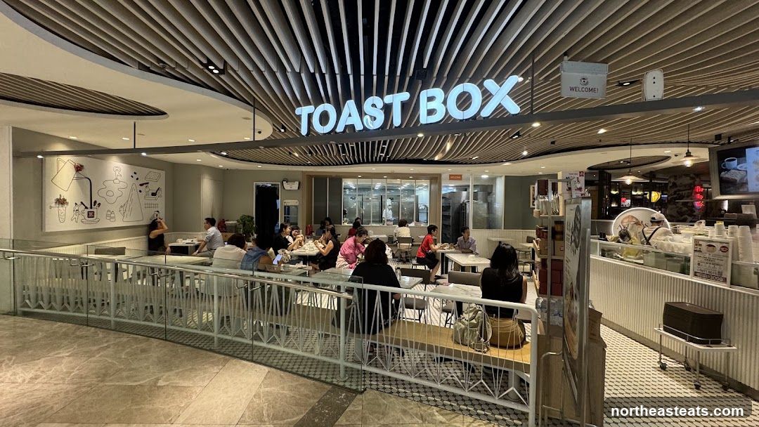 A wide shot of the interior of a Toast Box cafe. The cafe is filled with customers seated at tables. The brand name "TOAST BOX" is prominently displayed in large white letters above the counter. The cafe has a modern design with a slatted ceiling and a mural on the wall.