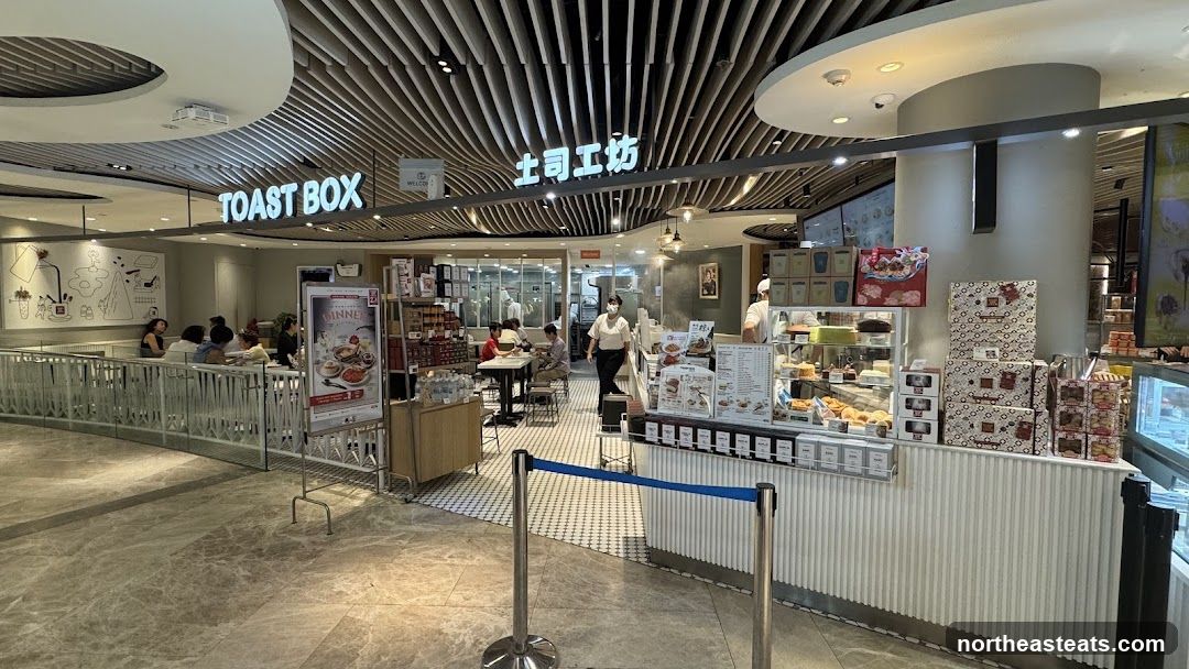 A view of the interior and exterior of a Toast Box cafe. The cafe has a modern design with wooden slatted ceilings and a white counter. There are tables and chairs for customers to sit at, and a display case filled with pastries and cakes. Staff members are visible behind the counter, preparing food and serving customers. A promotional poster is displayed near the entrance.