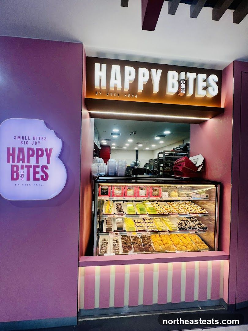 A brightly lit bakery storefront with a pink facade. The prominent signage reads "HAPPY BITES" in large white letters, with smaller text below indicating "BY SWEE HENG". To the left, a rounded illuminated sign also displays "HAPPY BITES" with the tagline "SMALL BITES BIG JOY". A glass display case showcases a variety of pastries and desserts arranged on multiple shelves. The lower part of the storefront features a pink and white striped design.
