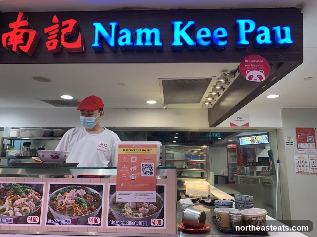 A storefront for Nam Kee Pau, a food establishment. A masked employee wearing a red cap and white shirt stands behind a counter. Above the counter, a neon sign reads "南記 Nam Kee Pau". Below the sign, there are three framed pictures of noodle dishes, each priced at $4.00. A Foodpanda sticker is visible on the right side of the sign. A sign on the counter indicates "Pau Order Here".
