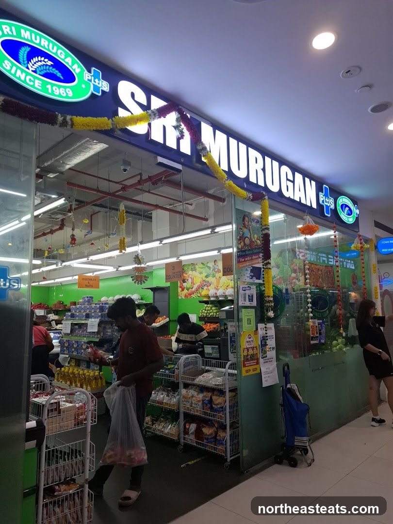 The exterior of a supermarket with a large illuminated sign that reads "SRI MURUGAN PLUS" and "SINCE 1969". The entrance is visible, with people shopping inside. Shelves stocked with various products are seen through the glass facade.