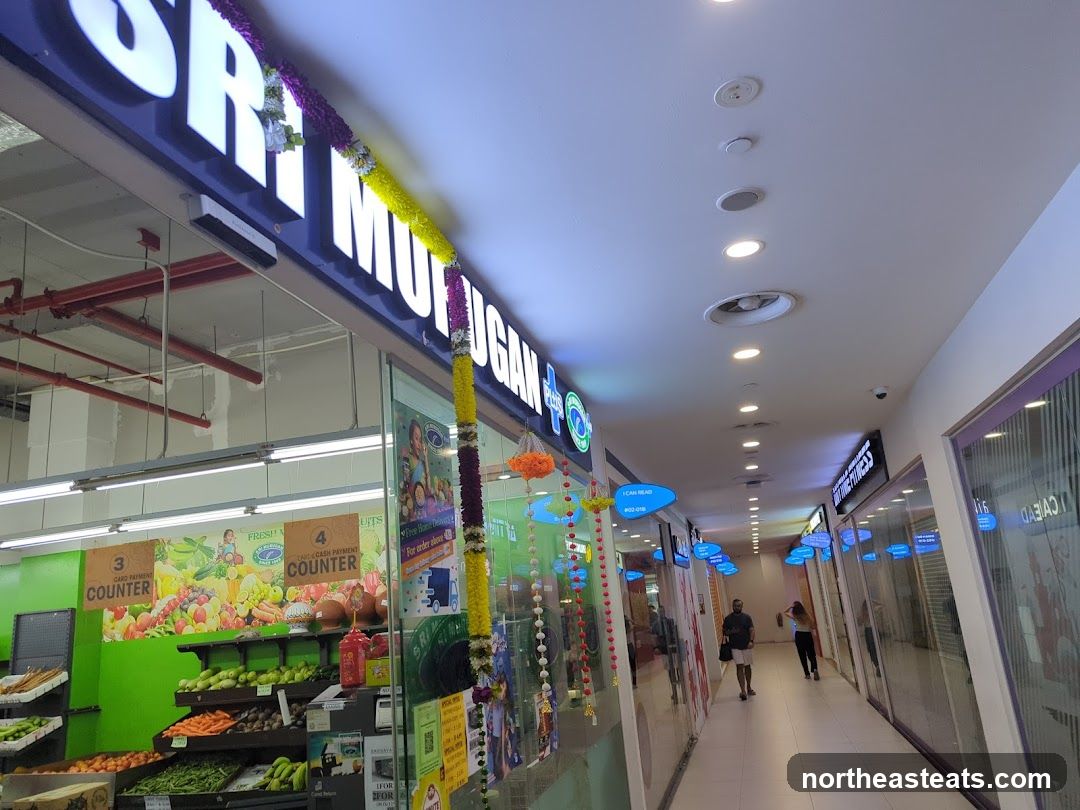 A brightly lit supermarket interior is visible through a glass storefront. The sign "SRI MUTUGAN" is prominently displayed above the entrance, adorned with colorful floral garlands. Inside, shelves are stocked with fresh produce, including fruits and vegetables. Several counters are labeled, with one indicating "CARD PAYMENT COUNTER" and another "PAYMENT COUNTER". The aisle extends into the distance, with other shops visible on either side.
