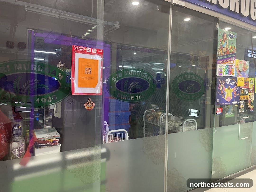 The image shows the storefront of Sri Murugan Plus, a store established in 1969. The store's name and logo are visible on the glass facade. Inside, shelves stocked with goods and shopping carts can be seen. Various promotional posters and flyers are displayed on the right side of the entrance, including one for "Happy Pongal" and another advertising free home delivery for orders above a certain amount. An orange notice with a QR code is also affixed to the glass.