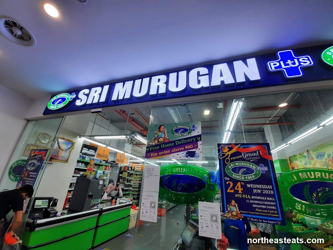 The exterior of a grocery store with a large illuminated sign that reads "SRI MURUGAN PLUS". The store has large glass windows displaying the interior, including shelves stocked with goods, checkout counters, and staff. Promotional posters are visible in the windows, announcing a grand opening and free home delivery.