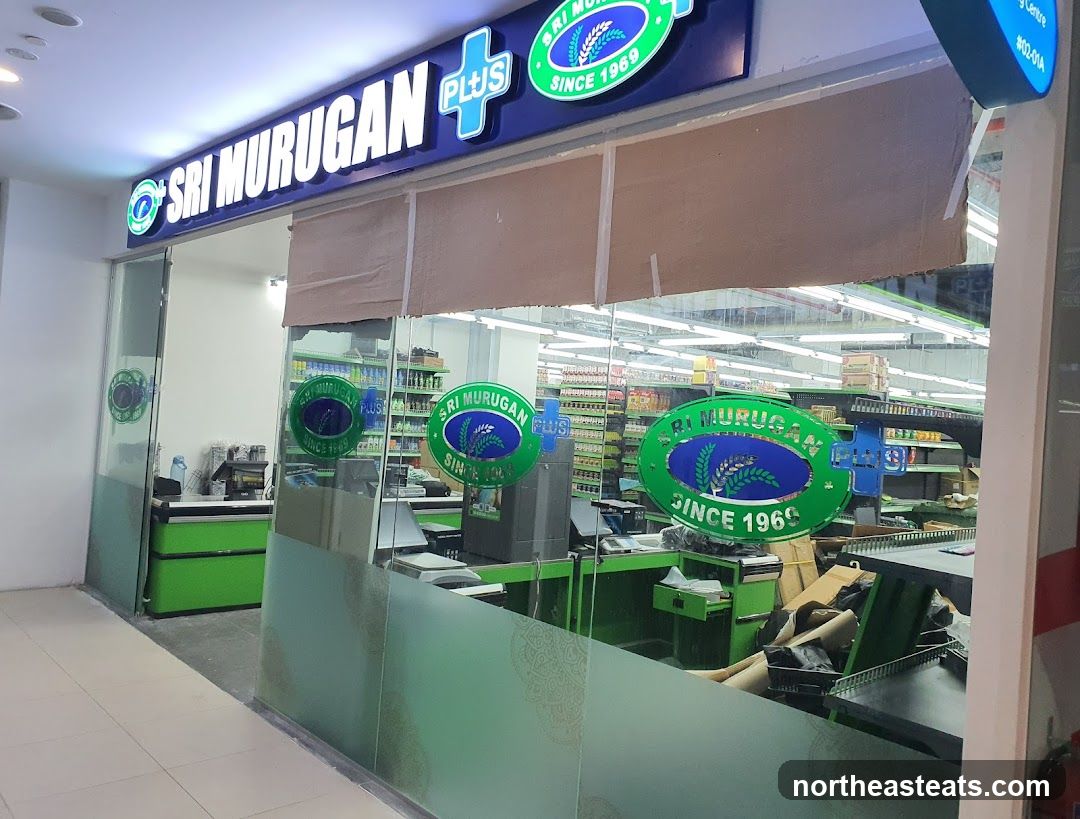 The exterior of a store called Sri Murugan Plus, with its name prominently displayed on a lit sign above the entrance. The store appears to be a supermarket, with shelves stocked with goods visible through the glass facade. Several circular green signs with the store's logo and "Since 1969" are also visible on the windows and inside.