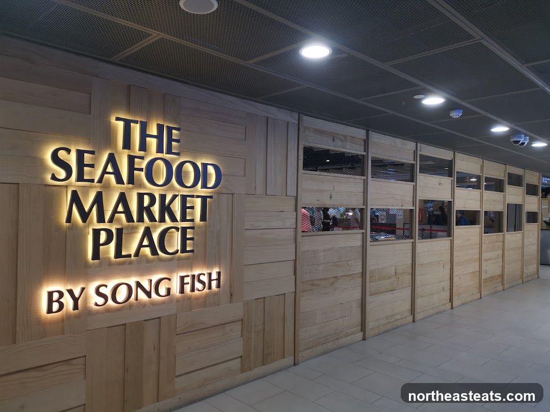 A brightly lit sign with the words "THE SEAFOOD MARKET PLACE BY SONG FISH" is mounted on a wooden paneled wall. The sign has illuminated lettering. To the right of the sign, the wall is divided into sections with wooden panels and mesh windows, revealing glimpses of the interior.