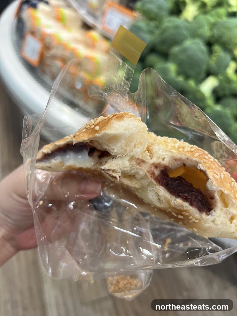 A close-up shot of a pastry, split in half, revealing a red bean filling and a yellow cheese-like substance. The pastry is coated with sesame seeds. It is held in a clear plastic bag. In the blurred background, there are shelves stocked with food items, including broccoli and what appear to be pre-packaged sandwiches or snacks.