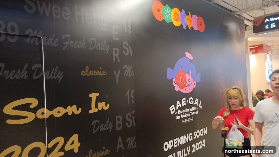 A black wall with text and graphics advertising a new bagel shop called BAE-GAL. The text reads "Soon In 2024" and "OPENING SOON IN JULY 2024". There is also a graphic of a character with bagels and the tagline "Bagels with an Asian Twist". People are walking by in the background.