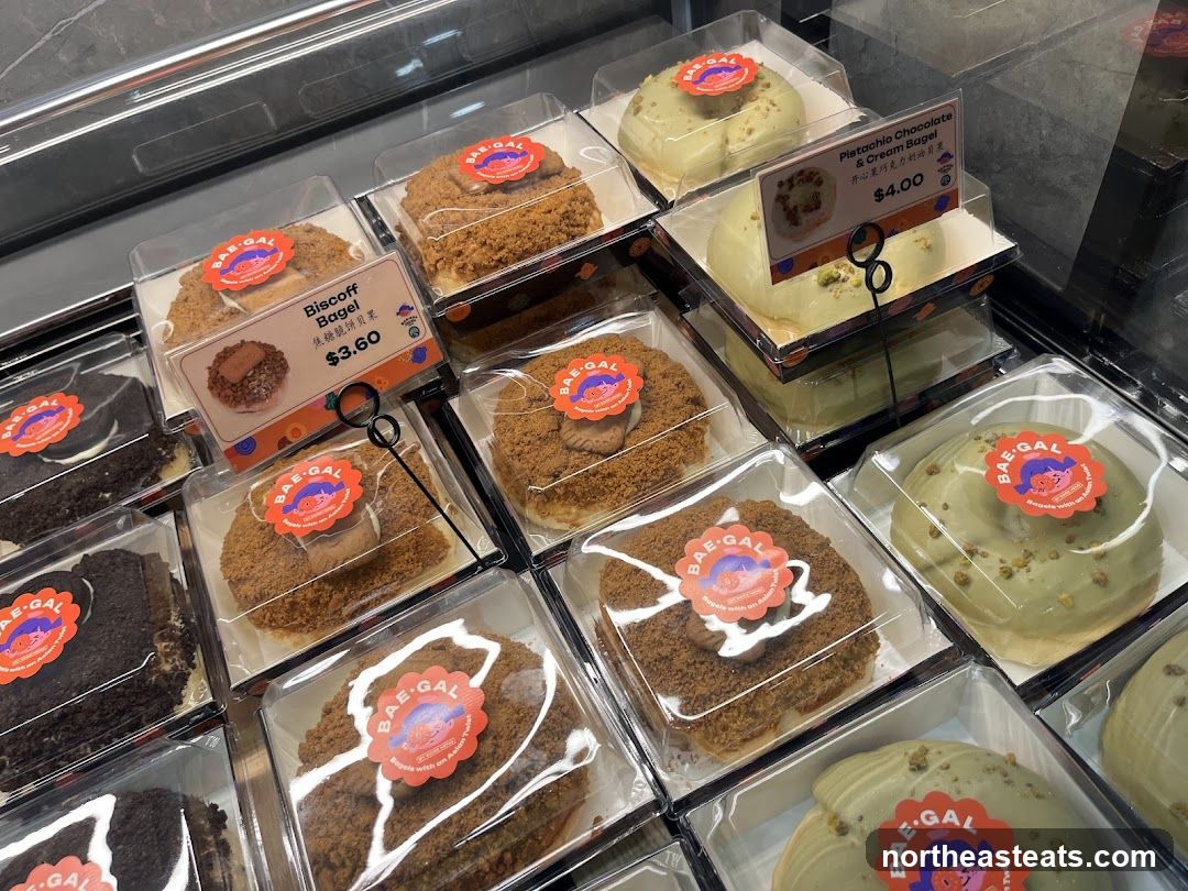 A display case filled with various types of bagels, each individually packaged. Visible flavors include Biscoff Bagel and Pistachio Chocolate & Cream Bagel. Each bagel has a circular sticker with the brand logo "BAE-GAL".