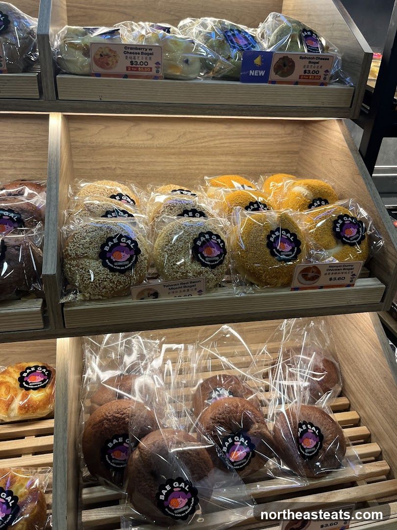 A display of various bagels in plastic packaging on wooden shelves. Some bagels are topped with sesame seeds, while others are plain or have a yellow coating. Labels indicate flavors such as Cranberry w Cheese, Spinach Cheese, and Taiwan Red Bean Mochi.