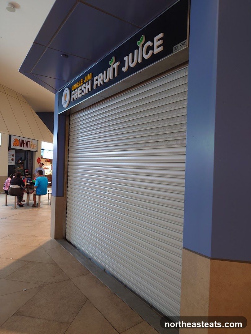 The exterior of a closed Uncle Jim Fresh Fruit Juice stall. The metal security shutter is down, covering the storefront. Above the shutter, a sign reads "UNCLE JIM FRESH FRUIT JUICE" with a small logo of a glass of juice. The stall is part of a larger indoor shopping area, with other shops and seating visible in the background.