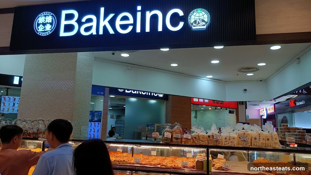 A brightly lit bakery storefront with a large illuminated sign that reads "Bakeinc" and features a Halal certification logo. Inside, display cases are filled with various baked goods, including bread and pastries. Customers are visible browsing the selection.