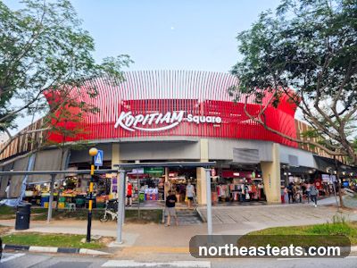 The exterior of a building with a prominent red and white striped facade. The words "Kopitiam square" are displayed in large white lettering on the red section. Below the signage, there are multiple storefronts with awnings, displaying various goods and people browsing. The area appears to be a market or food court.