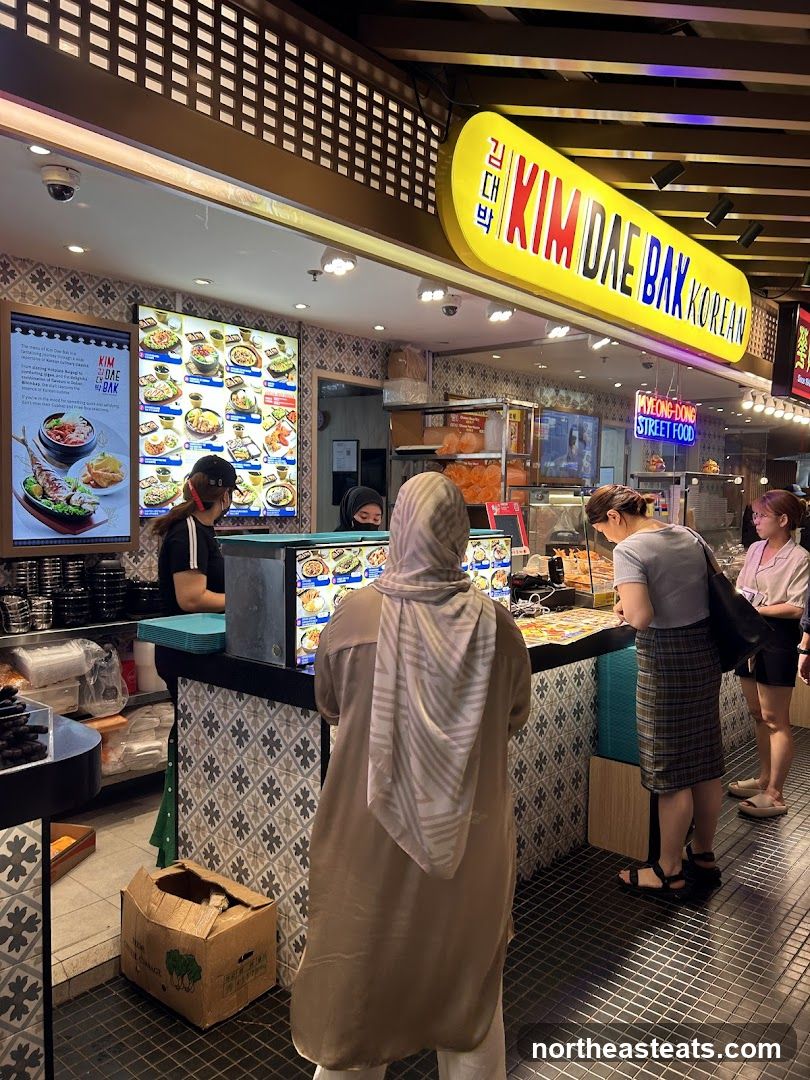 A bright yellow sign with red and blue lettering reads "KIM DAE BAK KOREAN". Below the sign, a food stall displays various Korean dishes on digital menus. Customers are seen ordering and waiting for their food.