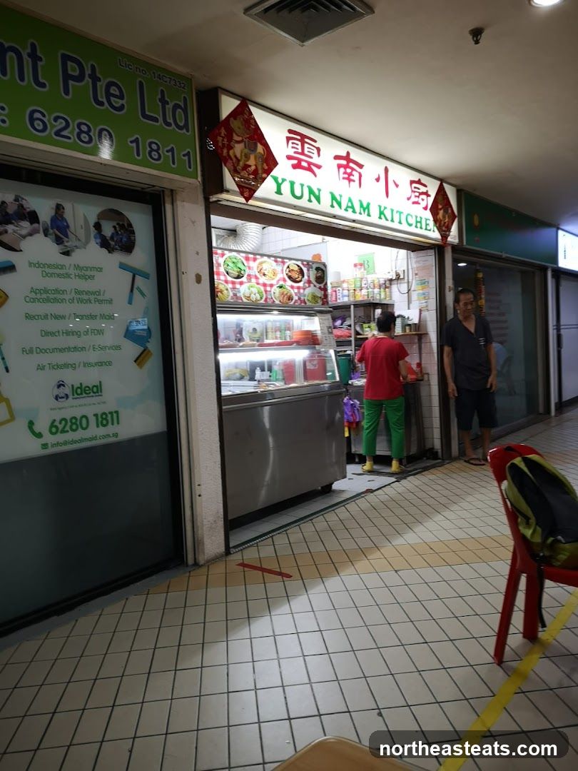 A food stall with a sign that reads "YUN NAM KITCHEN" in English and Chinese characters. There are pictures of food displayed above the counter. Two people are visible behind the counter, and another person is standing to the right. The stall is located in a mall or covered walkway.