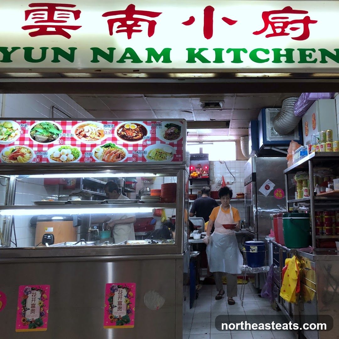 A food stall with a sign that reads "Yun Nam Kitchen" in English and Chinese characters. Below the sign is a menu board displaying various noodle dishes with prices. Inside the stall, staff members are preparing food.