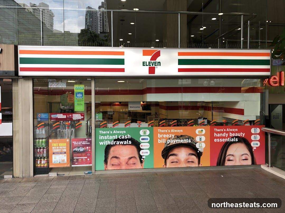 The exterior of a 7-Eleven convenience store. The prominent signage features the 7-Eleven logo and the brand's signature red, green, and orange stripes. The storefront has large glass windows displaying various promotional posters and advertisements. Inside, shelves stocked with products are visible.