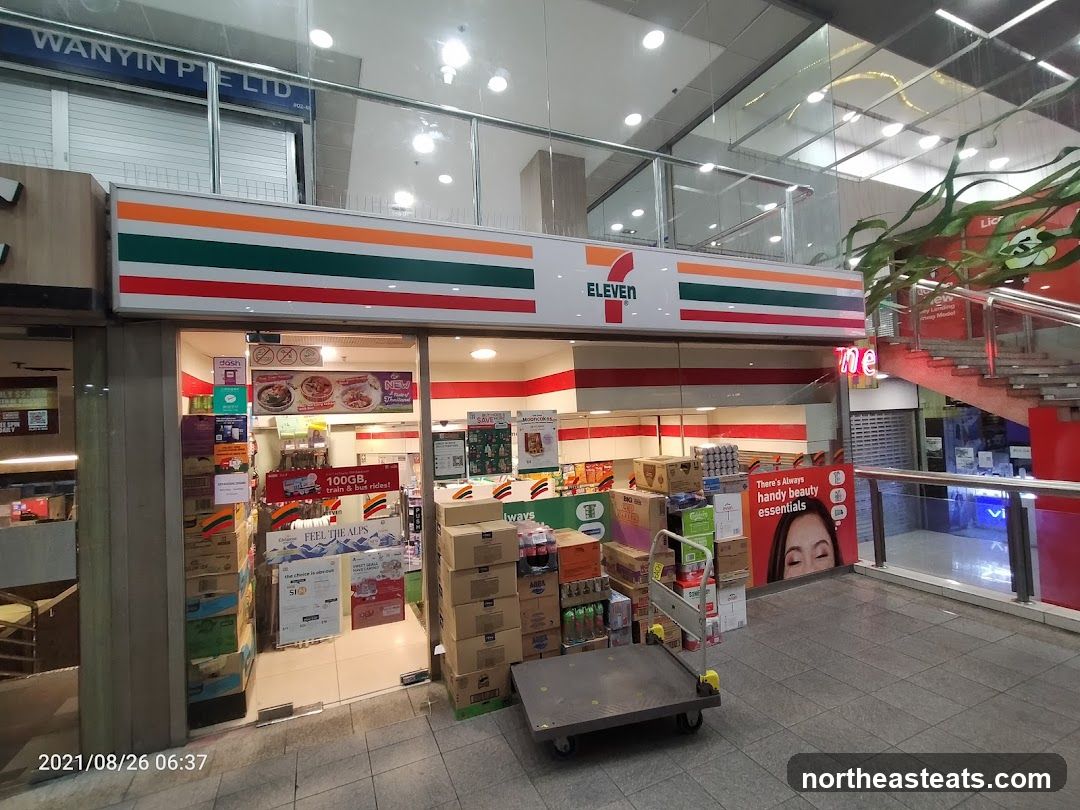 The exterior of a 7-Eleven convenience store with its signature red, green, and orange striped signage above the entrance. The store is stocked with various products, including boxes of drinks and promotional displays. A shopping cart is parked in front of the entrance.