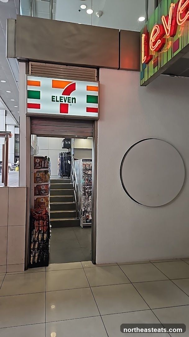 The image shows the entrance to a 7-Eleven convenience store. A prominent illuminated sign with the 7-Eleven logo is visible above the entrance. The store entrance is partially open, revealing a set of stairs leading down into the store. Shelves stocked with snacks and other merchandise are visible on either side of the entrance and on the stairs. A neon sign, partially visible on the right, appears to say "level" or similar.