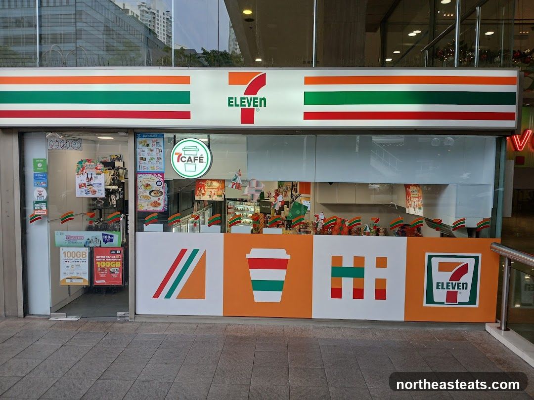 The exterior of a 7-Eleven convenience store. The store has a large illuminated sign with the 7-Eleven logo and colorful stripes. The storefront features large glass windows and doors, with various promotional posters and signage visible inside and on the glass. The lower part of the facade is decorated with colorful graphics, including a coffee cup icon and the 7-Eleven logo.