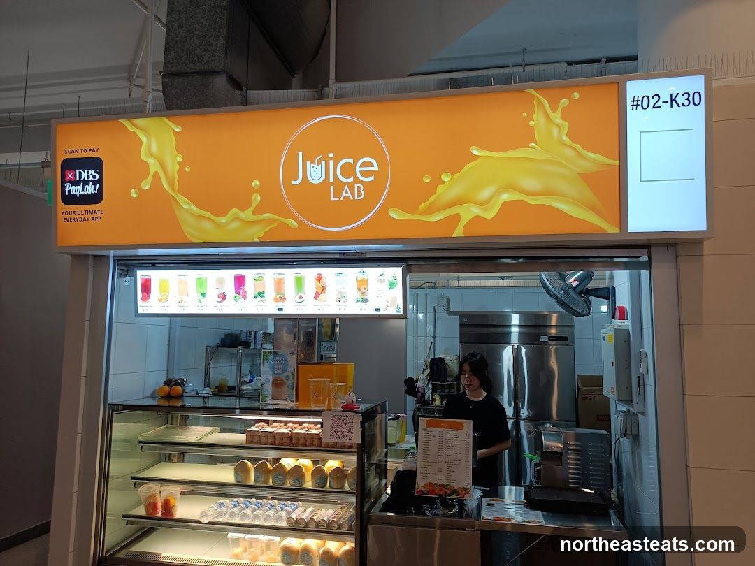 A juice bar with a bright orange sign featuring the logo "Juice Lab" and a splash of orange juice. Below the sign is a display case with various drinks and food items. A staff member is visible behind the counter.