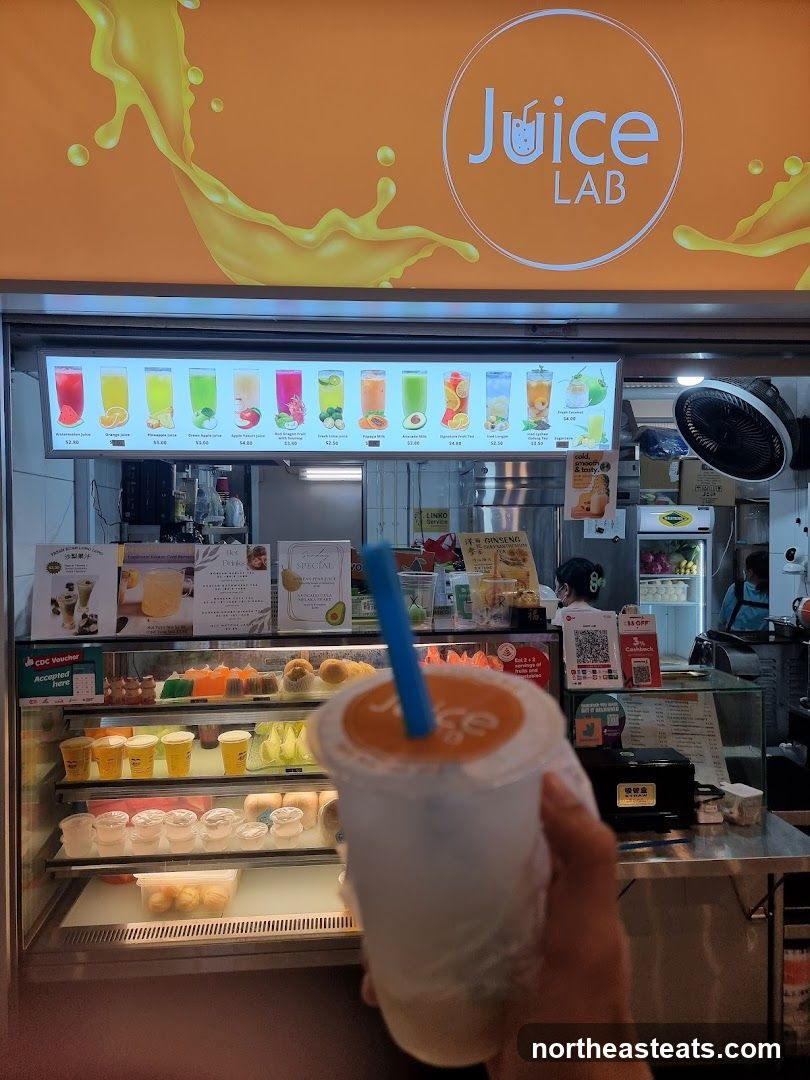 A storefront for a juice bar with a large illuminated menu board displaying various juices and their prices. Below the menu, a refrigerated display case shows an assortment of desserts and drinks. A hand holds a cup with the "Juice Lab" logo.