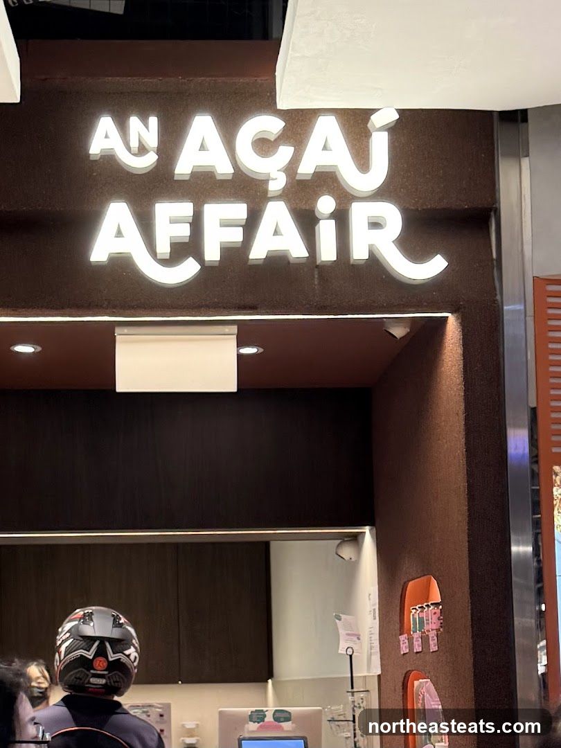 The image shows the illuminated signage for "AN AÇAI AFFAIR" above a dark brown facade. Below the sign, a counter area is visible with a person wearing a helmet, and some cabinets. To the right, there are some decorative orange elements on the wall with small cards attached.