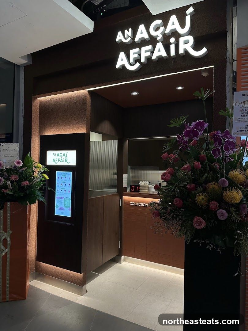 The exterior of an Açaí Affair shop. A large illuminated sign with the shop's name is visible above the entrance. To the left, a digital menu board displays various açaí bowl options. Floral arrangements are placed on either side of the entrance.