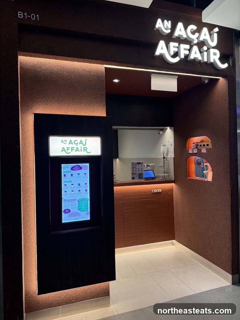 The exterior of a modern Açaí shop with a prominent illuminated sign displaying the business name. To the left, a digital menu board shows various açaí bowls and drinks. The counter area is visible inside, with an "ORDER" sign. The shop has a textured brown facade and is well-lit.