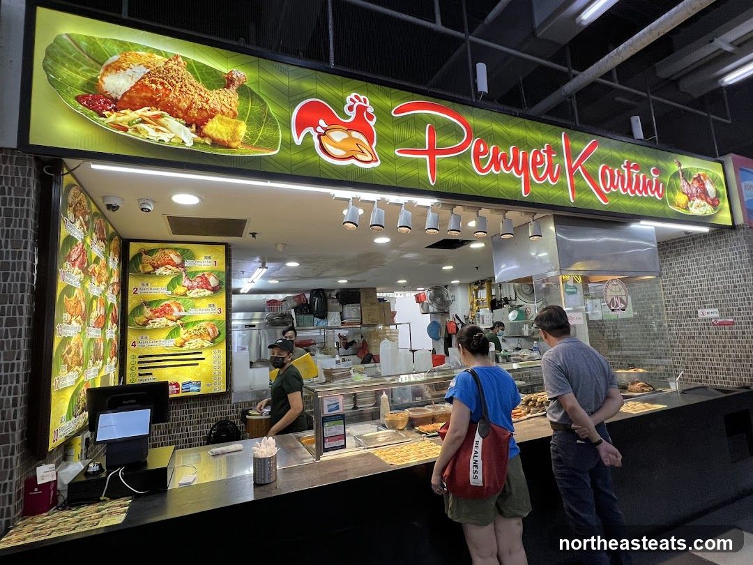 A food stall with a large illuminated sign displaying the name "Penyet Kartini" and images of food. There are menu boards on the left side of the stall, showing various chicken dishes with prices. Customers are seen ordering at the counter. The stall appears to be in a food court or market setting.