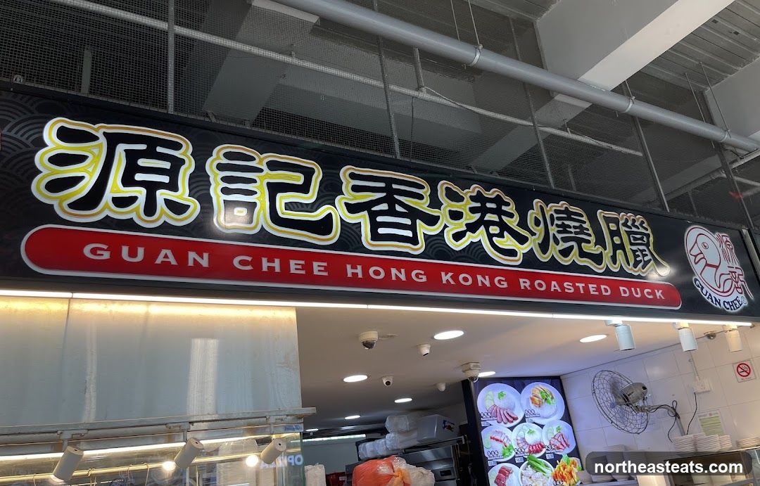 A black sign with yellow and white Chinese characters and the English text "GUAN CHEE HONG KONG ROASTED DUCK" is displayed above a food stall. A cartoon duck logo is visible on the right side of the sign. Below the sign, a menu board displays images of roasted duck and other dishes with prices.