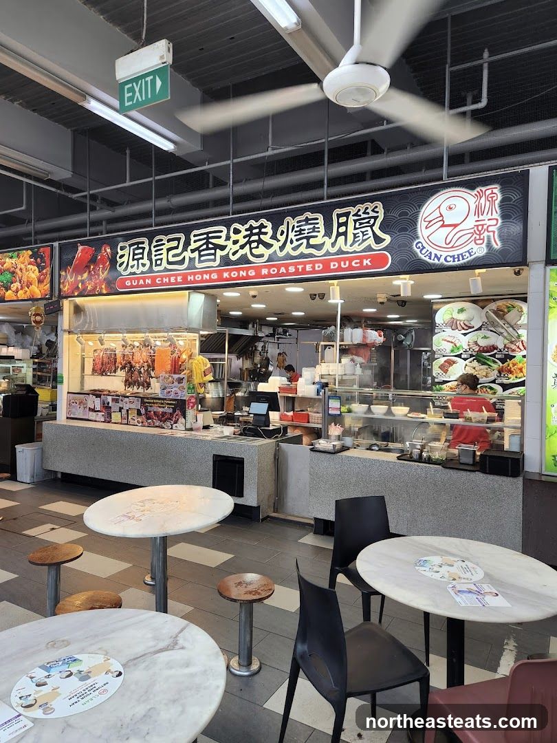 A food stall with a prominent sign displaying "Guan Chee Hong Kong Roasted Duck" in both Chinese and English. Roasted ducks and other meats are displayed in a glass case. There are tables and chairs in front of the stall.