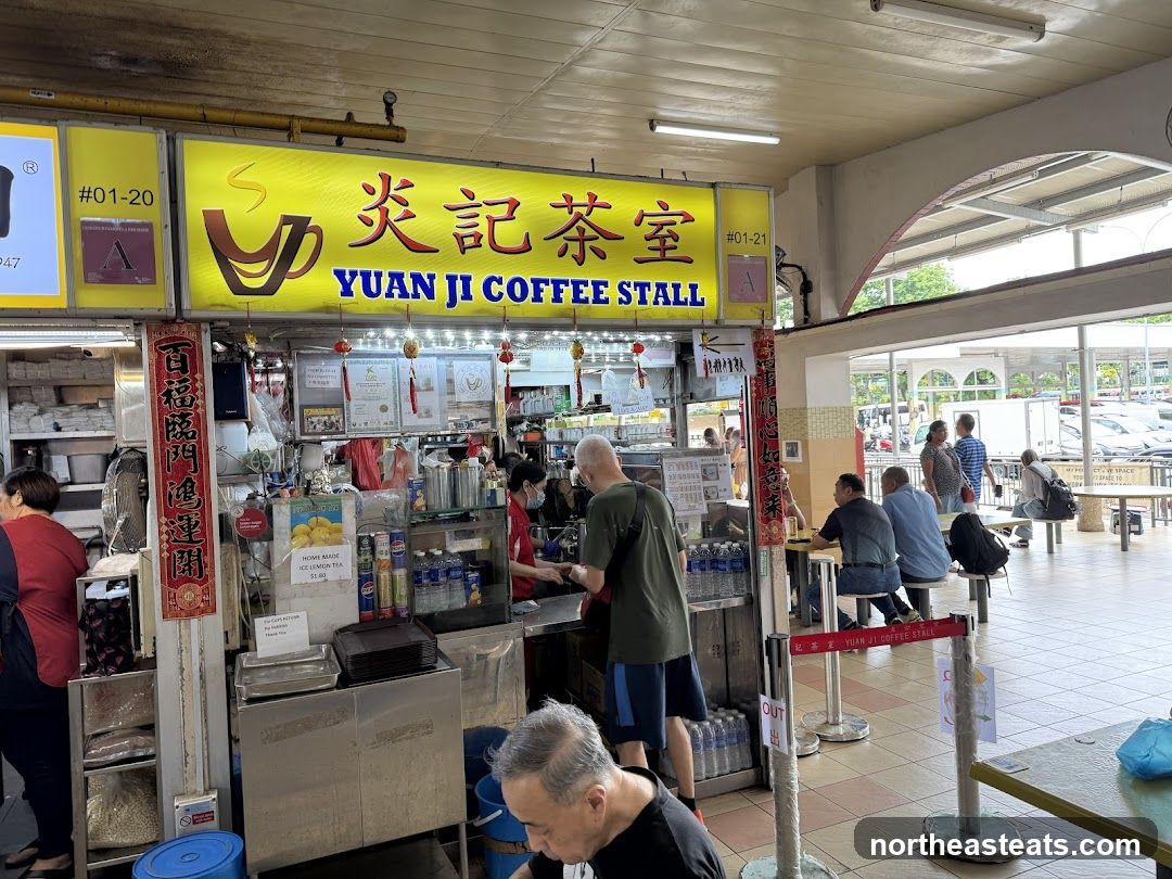 A brightly lit coffee stall with a large yellow sign displaying the name "YUAN JI COFFEE STALL" in English and Chinese characters. The stall is busy with customers and staff. There are various drinks and food items displayed. The stall is located in an open-air food court or hawker center.