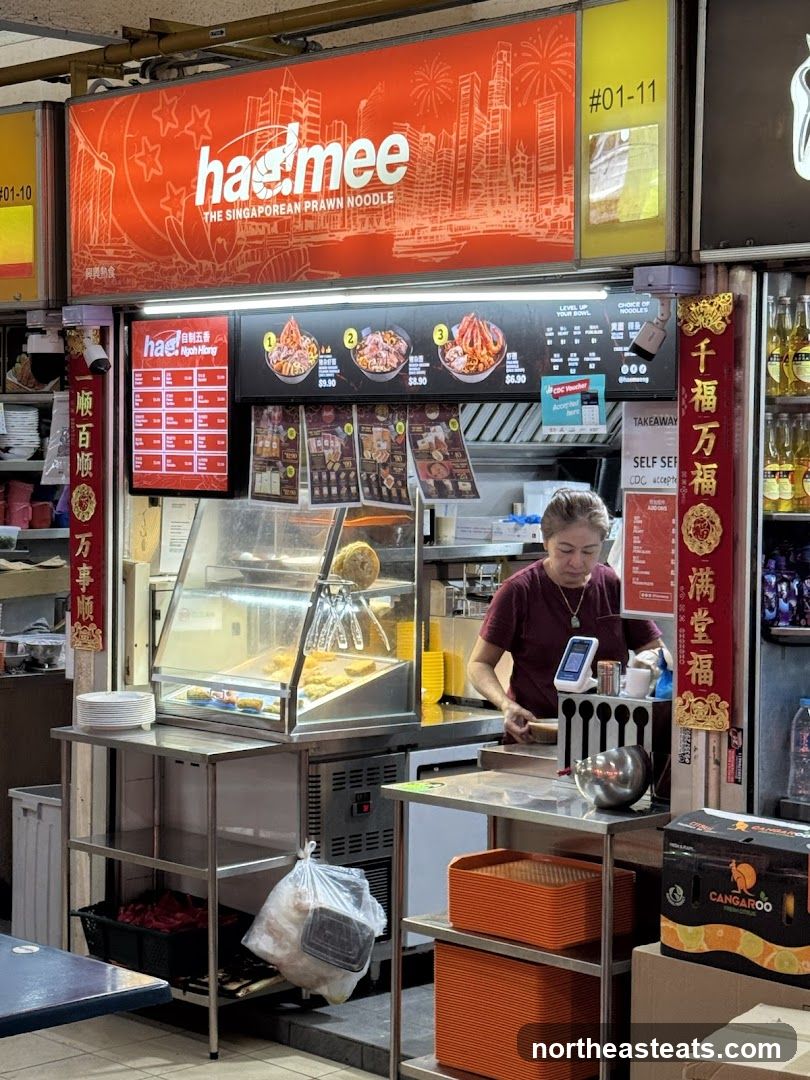 A food stall with a large orange sign displaying the business name and a cityscape graphic. Below the sign, a menu board shows various prawn noodle dishes with prices. A staff member is visible behind the counter, preparing food.