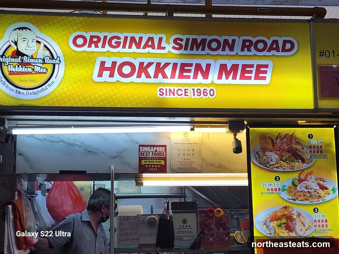 A bright yellow sign with red lettering reads "ORIGINAL SIMON ROAD HOKKIEN MEE SINCE 1960". To the left is a cartoon image of a man giving a thumbs up. Below the sign is a menu board displaying three dishes with prices and descriptions. A man wearing a mask is visible behind the counter.
