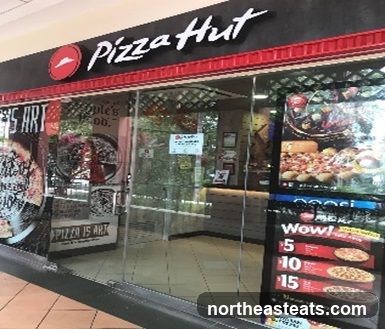 The exterior of a Pizza Hut restaurant with large glass doors and windows. The Pizza Hut logo and name are visible above the entrance. Inside, a counter and seating area can be seen. A promotional display with pizza images and prices is located to the right of the entrance.