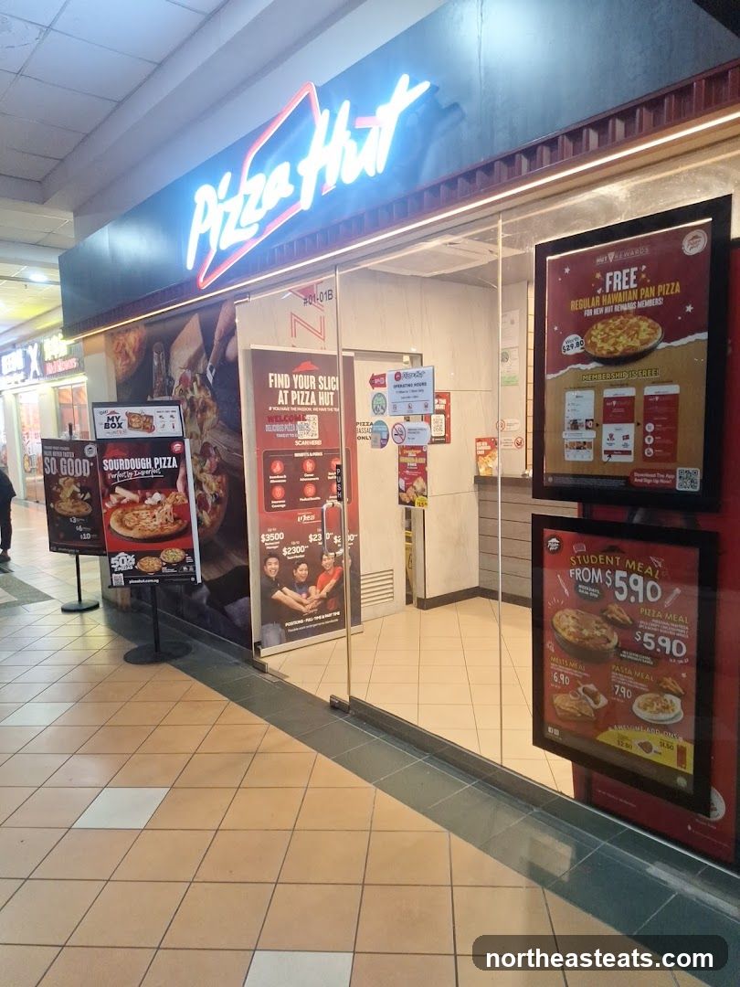 The exterior of a Pizza Hut restaurant with a large neon sign above the entrance. There are several promotional posters displayed on the glass doors and on stands outside the entrance.