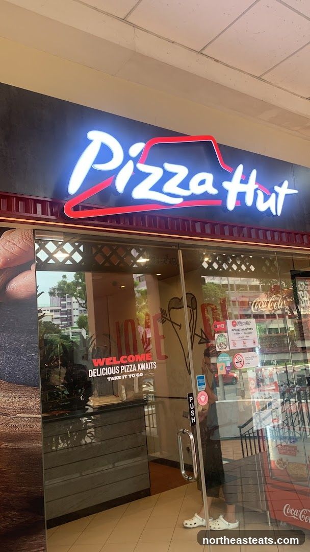 The exterior of a Pizza Hut restaurant with a large illuminated sign above the entrance. The glass doors have a "Welcome, Delicious Pizza Awaits, Take It To Go" decal.