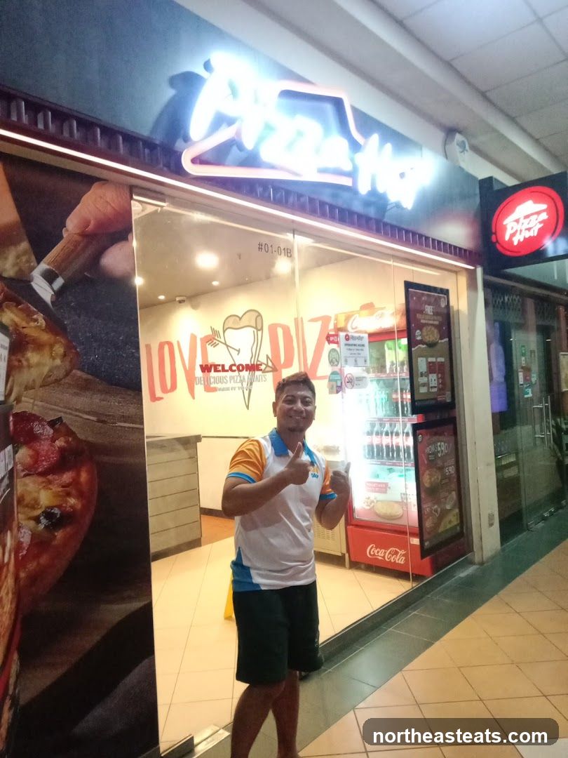 A man gives a thumbs up in front of a Pizza Hut storefront. The store has a large neon Pizza Hut sign above the entrance and a smaller red Pizza Hut logo sign to the right. The glass doors and windows display pizza imagery and promotional materials. Inside, a Coca-Cola refrigerator is visible.