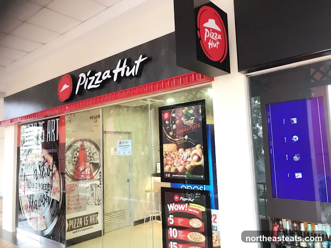 The exterior of a Pizza Hut restaurant with prominent signage. The main sign above the entrance reads "Pizza Hut" in white lettering on a dark background, with the iconic red Pizza Hut logo to the left. A smaller, hanging sign with the Pizza Hut logo is also visible. The storefront features large glass windows with various posters and graphics, including one advertising "Loaded Puffs" and another with the slogan "Pizza is Art". A digital screen is mounted to the right of the entrance.