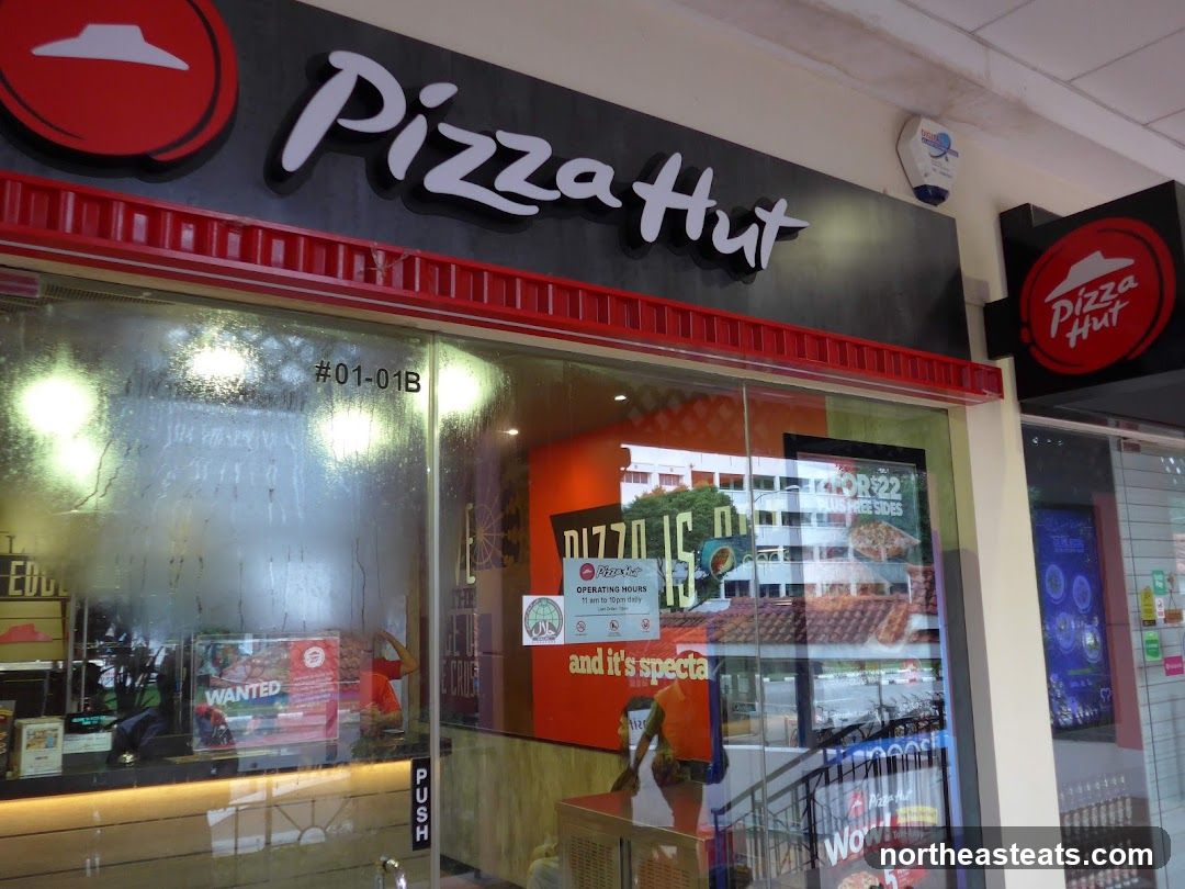 The exterior of a Pizza Hut restaurant with prominent signage. The main sign above the entrance reads "Pizza Hut" in large white lettering with a red accent. A circular Pizza Hut logo is visible to the right of the main sign. The storefront features large glass windows, through which some interior elements and promotional materials are visible. A "PUSH" sign is on the glass door.