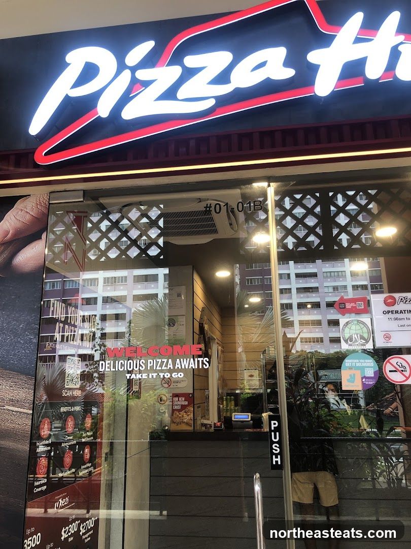 The exterior of a Pizza Hut restaurant. A large illuminated sign with the Pizza Hut logo is visible above the entrance. The glass doors have various stickers and signage, including a "Welcome, Delicious Pizza Awaits" message and information about delivery.