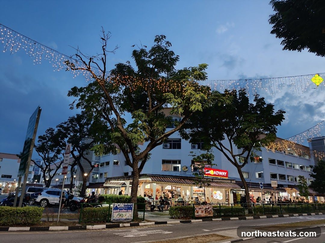 A row of shops with awnings and outdoor seating, illuminated by string lights. The sign for "BROADWAY" is visible in red neon letters above one of the establishments. People are seated at tables outside the shops. Trees with decorative lights are in the foreground.