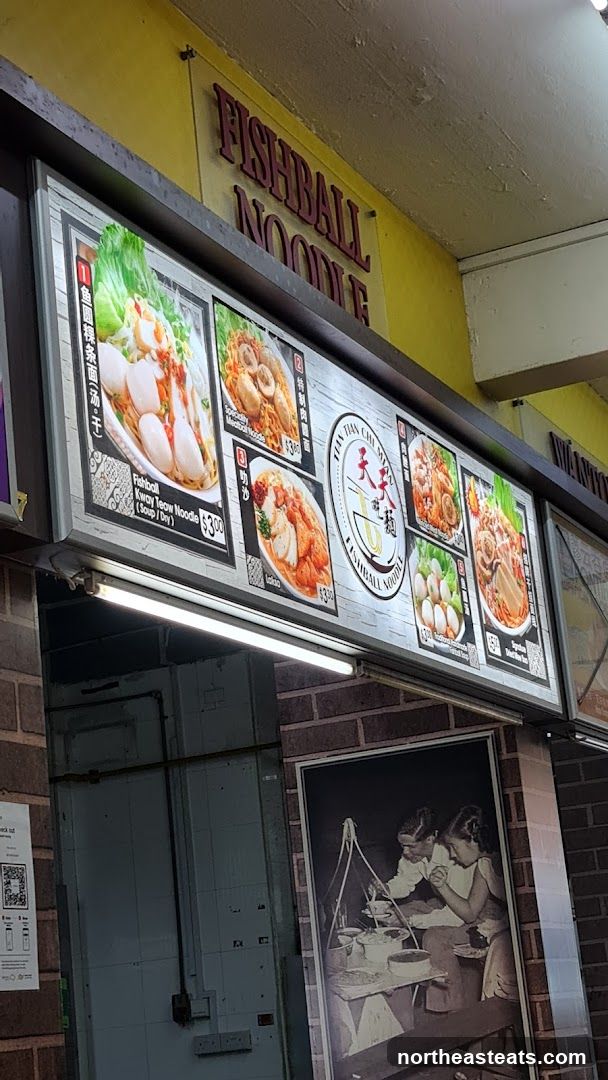 A brightly lit sign displays various noodle dishes with prices. The sign features images of different noodle bowls, including fishball kway teow noodle and laksa. The shop's name, "FISHBALL NOODLE", is prominently displayed in red lettering above the illuminated menu. Below the menu, a black and white framed picture shows a couple preparing food.