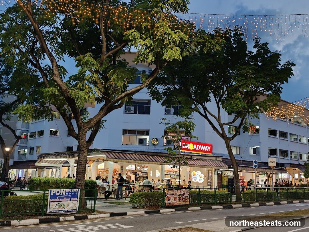 A row of food stalls with awnings and outdoor seating is visible under a building adorned with string lights. The "BROADWAY" sign is prominently displayed. People are seated at tables, and some are ordering food. A sign for "PON MINI MART" is also visible.