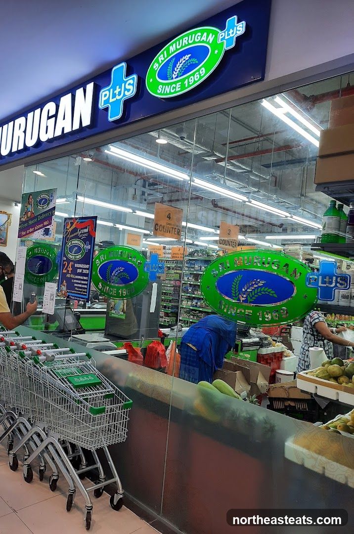 A wide shot of the entrance to a grocery store called Sri Murugan Plus. The store's name is prominently displayed on a large blue sign above the entrance, with a green oval logo featuring wheat stalks and the text "SRI MURUGAN PLUS SINCE 1969". Inside, shopping carts are lined up, and several employees are visible behind the counter, attending to customers and stocking shelves. Promotional posters are displayed on the glass windows.