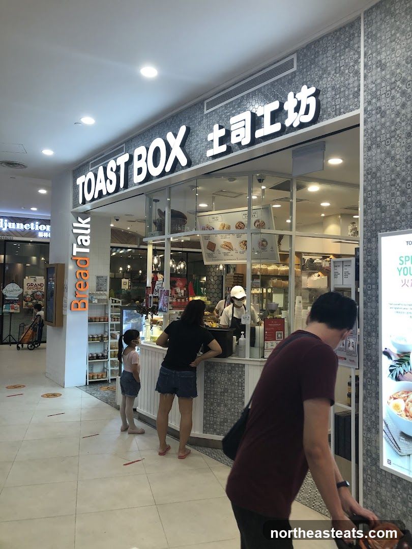 A view of the Toast Box storefront in a mall. The signage "TOAST BOX" is visible above the glass windows. Inside, staff members are serving customers. There are also displays of food items and menus visible through the windows.